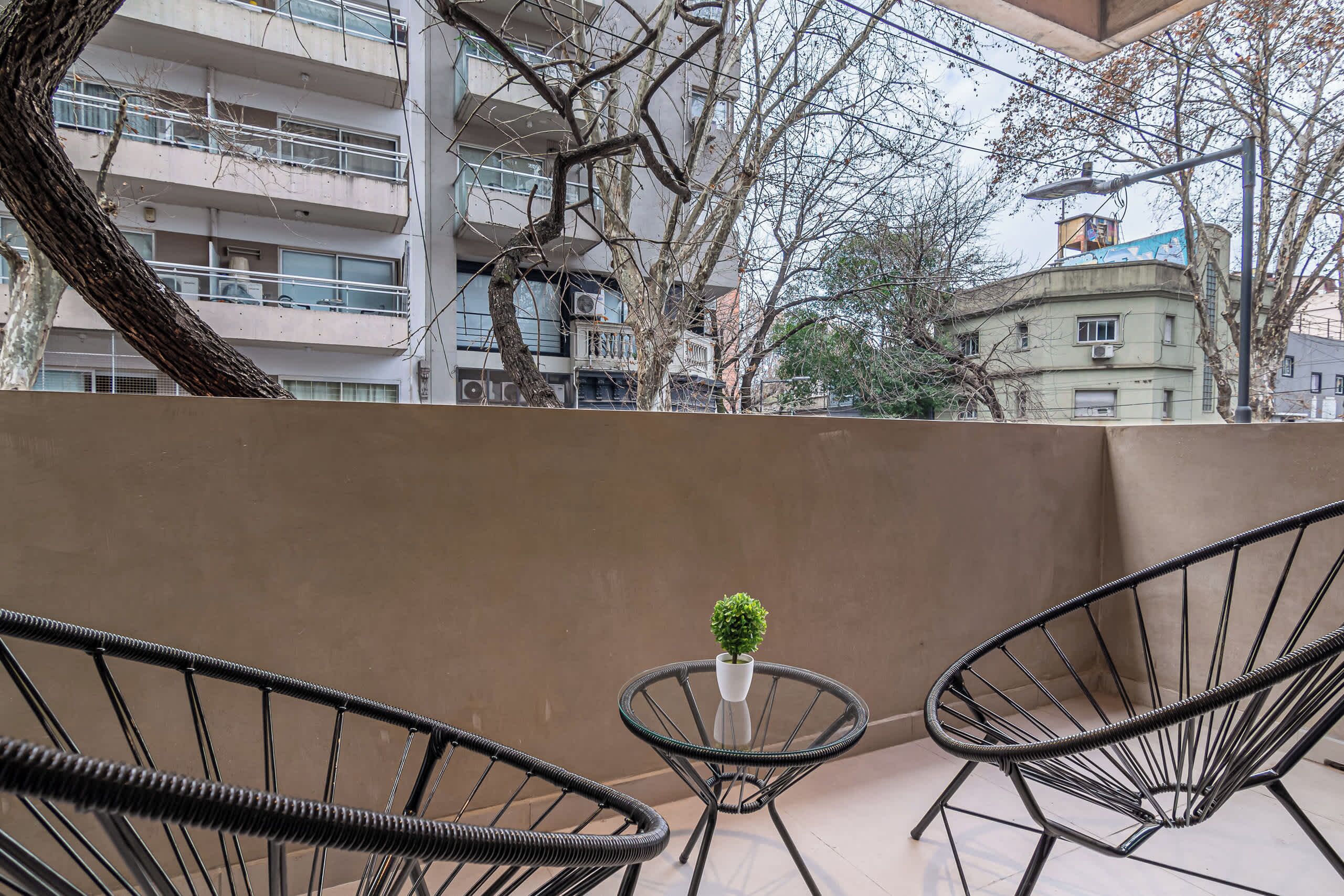 Un balcon avec deux chaises en métal, une petite table sur laquelle est posée une plante en pot, et une vue sur un immeuble à plusieurs étages doté de balcons.