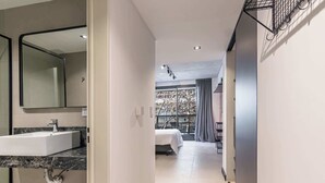 Een moderne hotelkamer met een groot raam, een bed, een badkamer met wastafel en spiegel, en een gang naar een andere kamer.