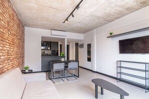 Ein modernes Wohnzimmer mit Backsteinwand, einer Couch, einem Essbereich mit Stühlen, einer Bank, einem Fernseher und einer Küche im Hintergrund.