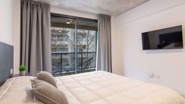 Un dormitorio con una cama grande, un televisor montado en la pared y un ventanal con cortinas.
