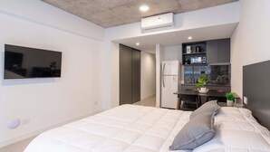 Un dormitorio moderno con una cama grande, un televisor montado en la pared, un escritorio empotrado con refrigerador y una pequeña planta en maceta.