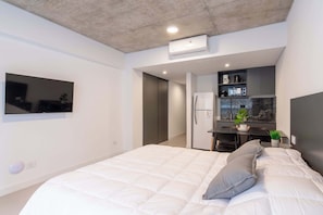 A modern bedroom with a large bed, a TV mounted on the wall, a built-in desk with a refrigerator, and a small potted plant. - Soho Village en Palermo by Magno | 501 (Buenos Aires)