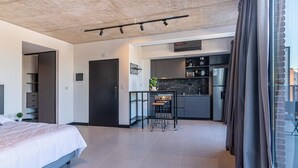 Een moderne hotelkamer met een bed, een kitchenette, een klein zitehoekje en een groot raam met gordijnen.