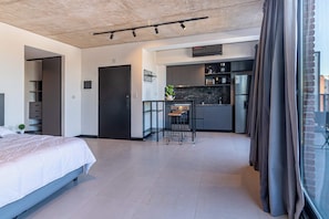 Ein modernes Hotelzimmer mit Bett, einer Küchenzeile, einer kleinen Essecke und einem großen Fenster mit Vorhängen.
