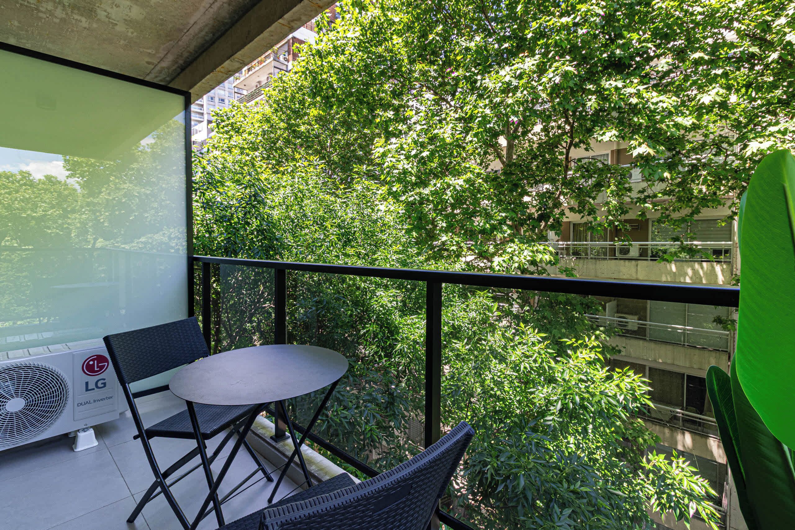 Un balcon avec une table et des chaises, un ventilateur, et une vue sur les arbres et les bâtiments.