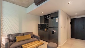 Ein modernes Wohnzimmer mit Sofa, Couchtisch, Küchenzeile inklusive Kühlschrank und einer dunklen Tür.