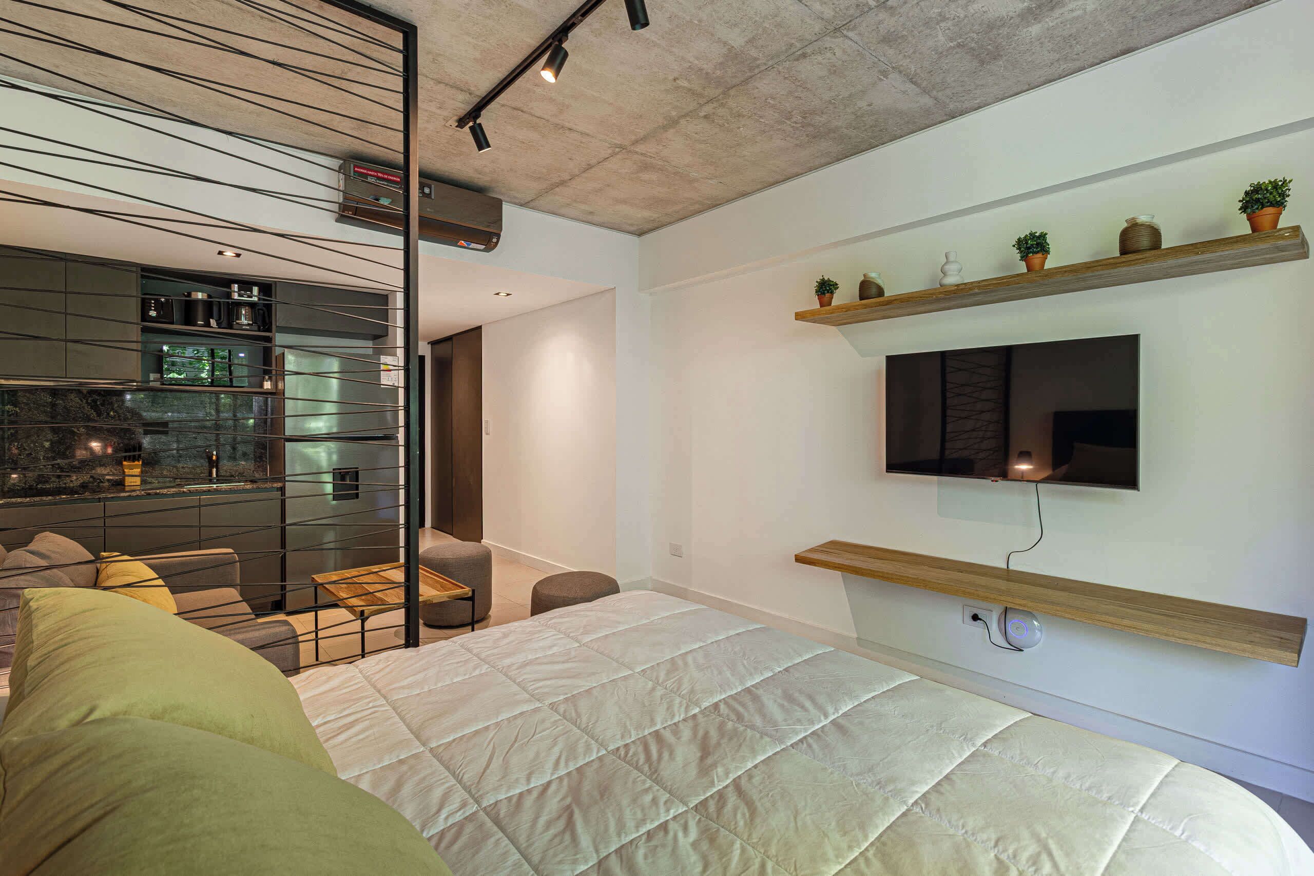 Ein modernes Schlafzimmer mit einem großen Bett, einem an der Wand montierten Fernseher und einer durch eine Glastrennwand sichtbaren Küchenzeile.