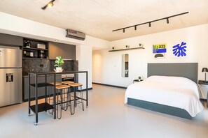 Ein modernes Hotelzimmer mit einer Küchenzeile, einem Bett, einem kleinen Tisch mit Hocker und einem Spiegel.