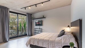 Un dormitorio moderno con una cama grande, una mesita pequeña, un televisor y un balcón con plantas.