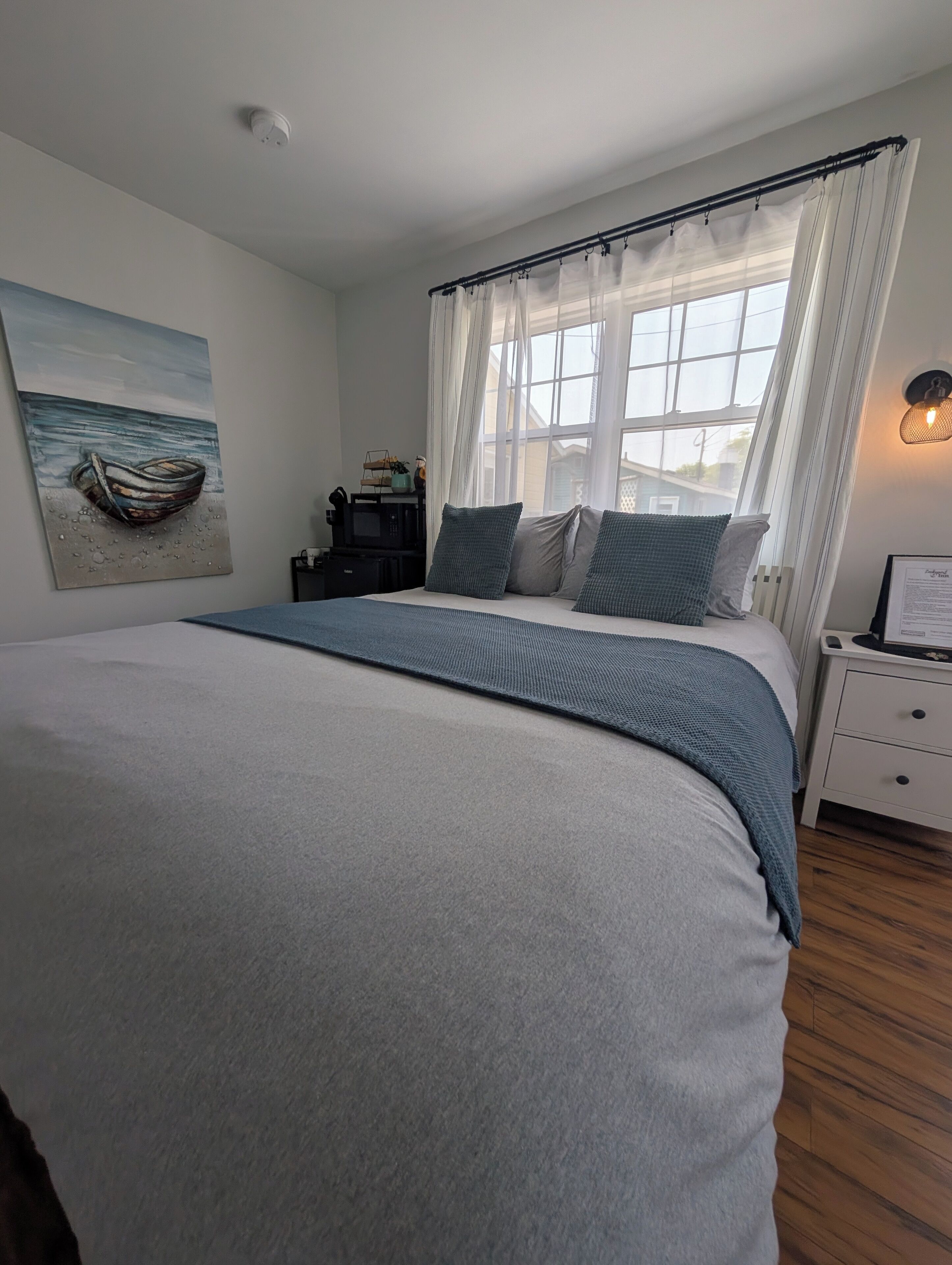 A bedroom with a bed, a nightstand, a window with curtains, a wall-mounted light, and a painting.