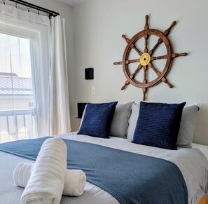 A bed with a blue and white bedspread, two pillows, a rolled white towel, and a wooden ship's wheel on the wall.