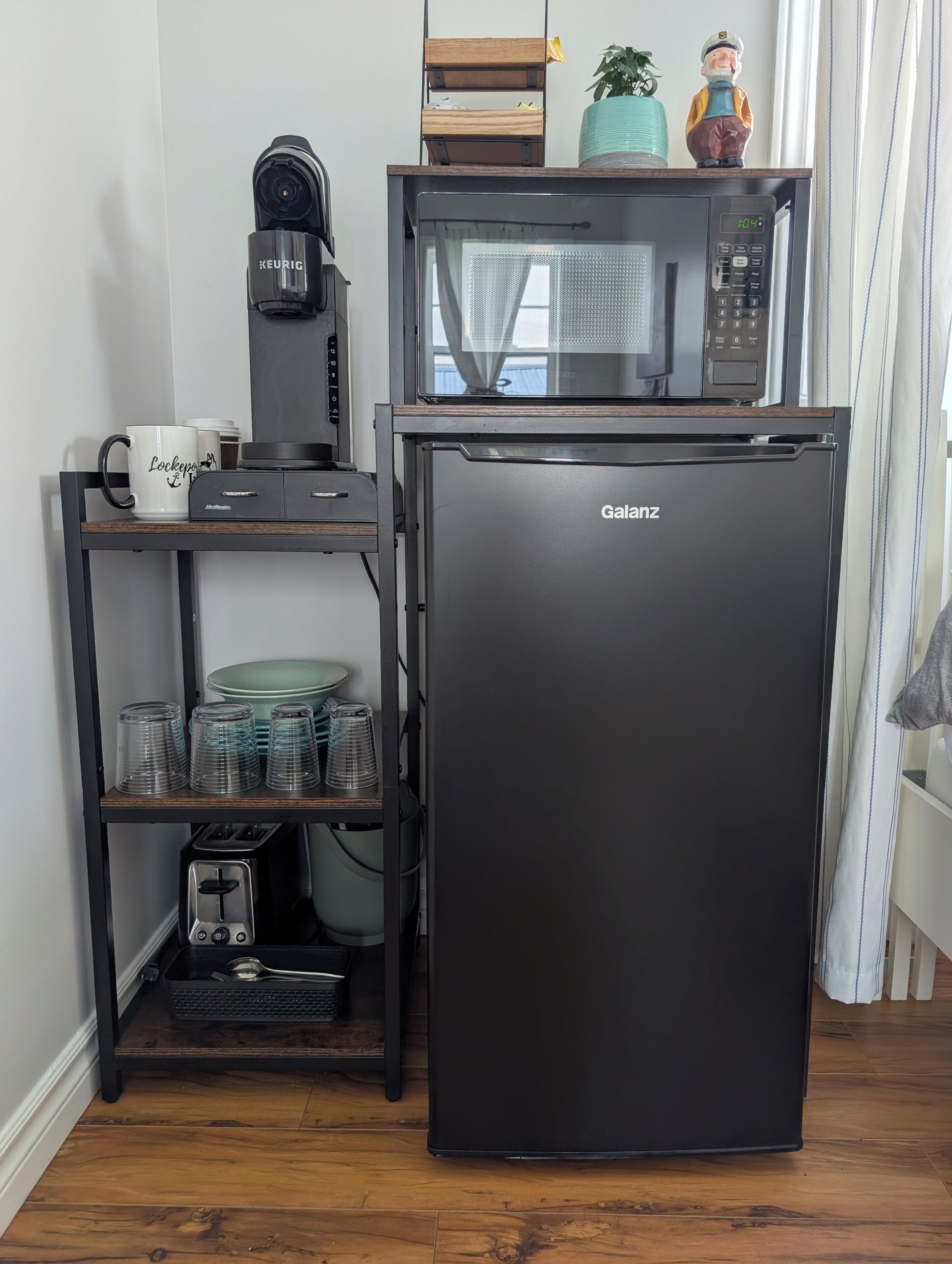 Una kitchenette compacta con refrigerador Galanz, cafetera Keurig, microondas y una taza de café sobre una repisa.