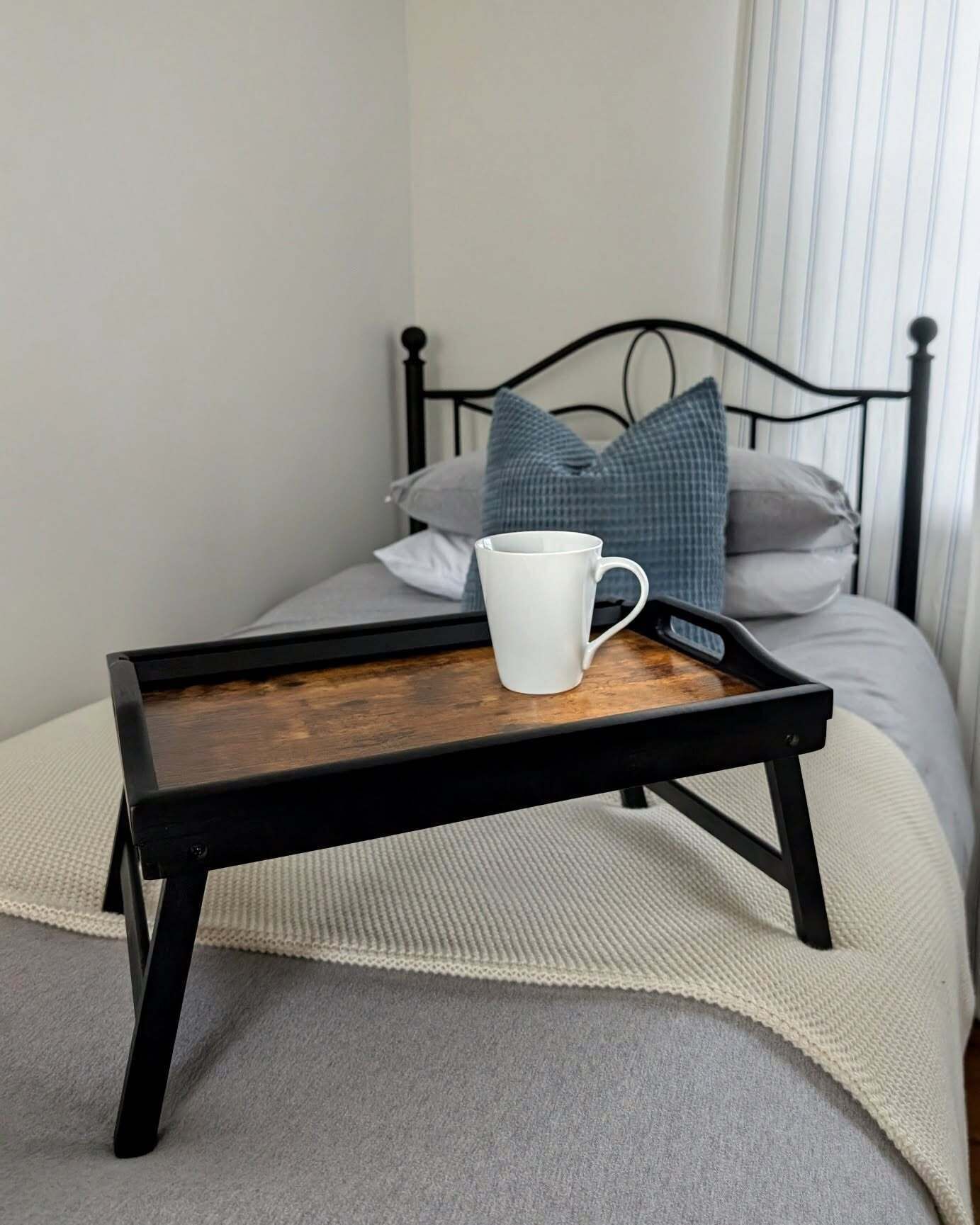 A bed with a black metal frame, a white pillow, a blue pillow, and a white mug on a wooden tray.