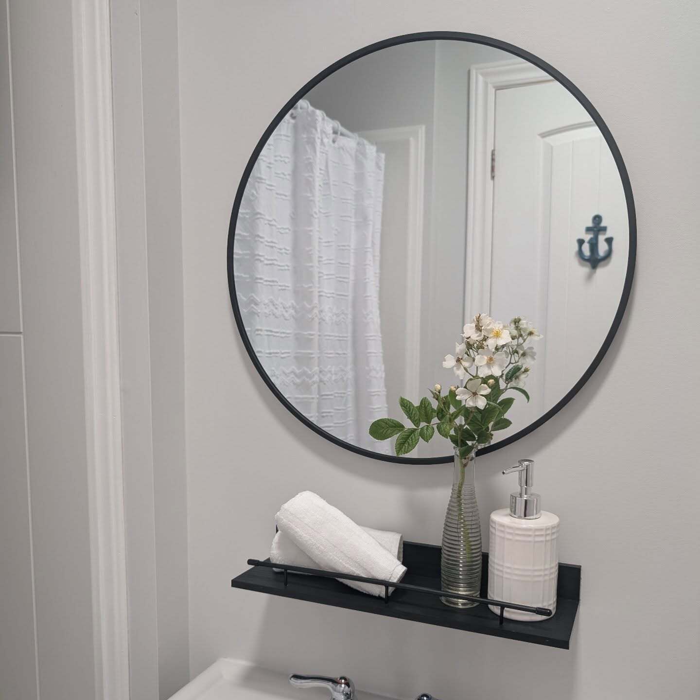 Ein Badezimmer mit rundem Spiegel, einem weißen Handtuch, einer Glasvase mit Blumen und einem weißen Toilettenpapierhalter.