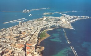 Une vue aérienne d’une ville côtière, avec un port imposant, une structure fortifiée et un mélange de bâtiments historiques et modernes.