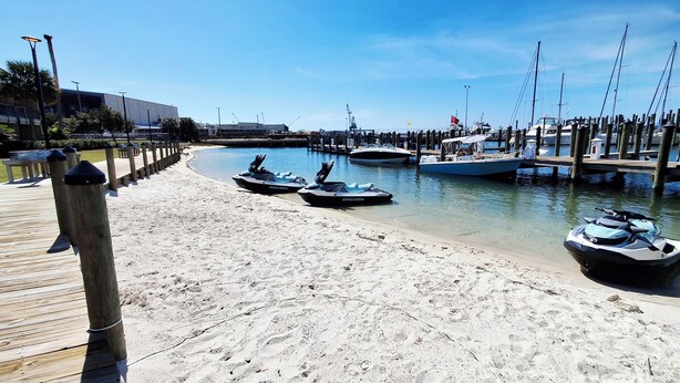 En sandstrand med jetski, en træbrygge og både i lysthavnen.