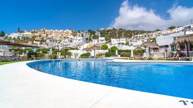 Une piscine avec des chaises longues, un ciel dégagé et, au loin, une colline parsemée de bâtiments.