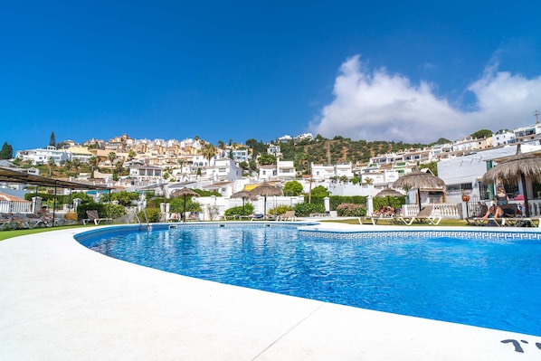A swimming pool with lounge chairs, a clear sky, and a hillside of buildings in the background. - Tropicana 6C by Casasol (Nerja)