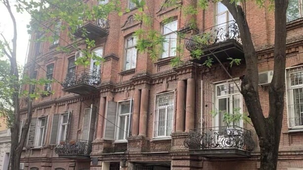 Un edificio de ladrillo de varios pisos con balcones, un coche estacionado al frente y un árbol en primer plano.