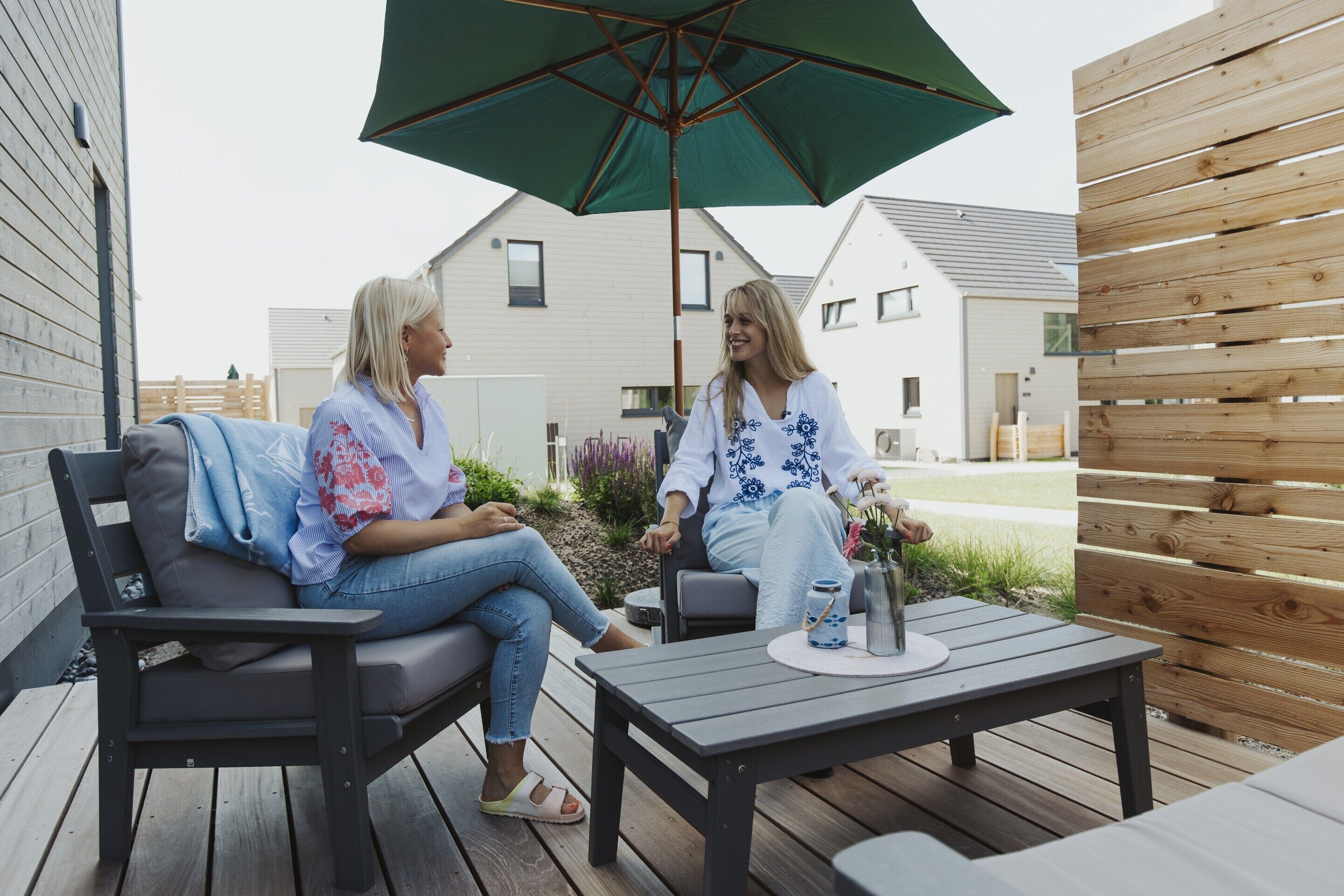 Two people sitting on a wooden deck with a table and a green umbrella.