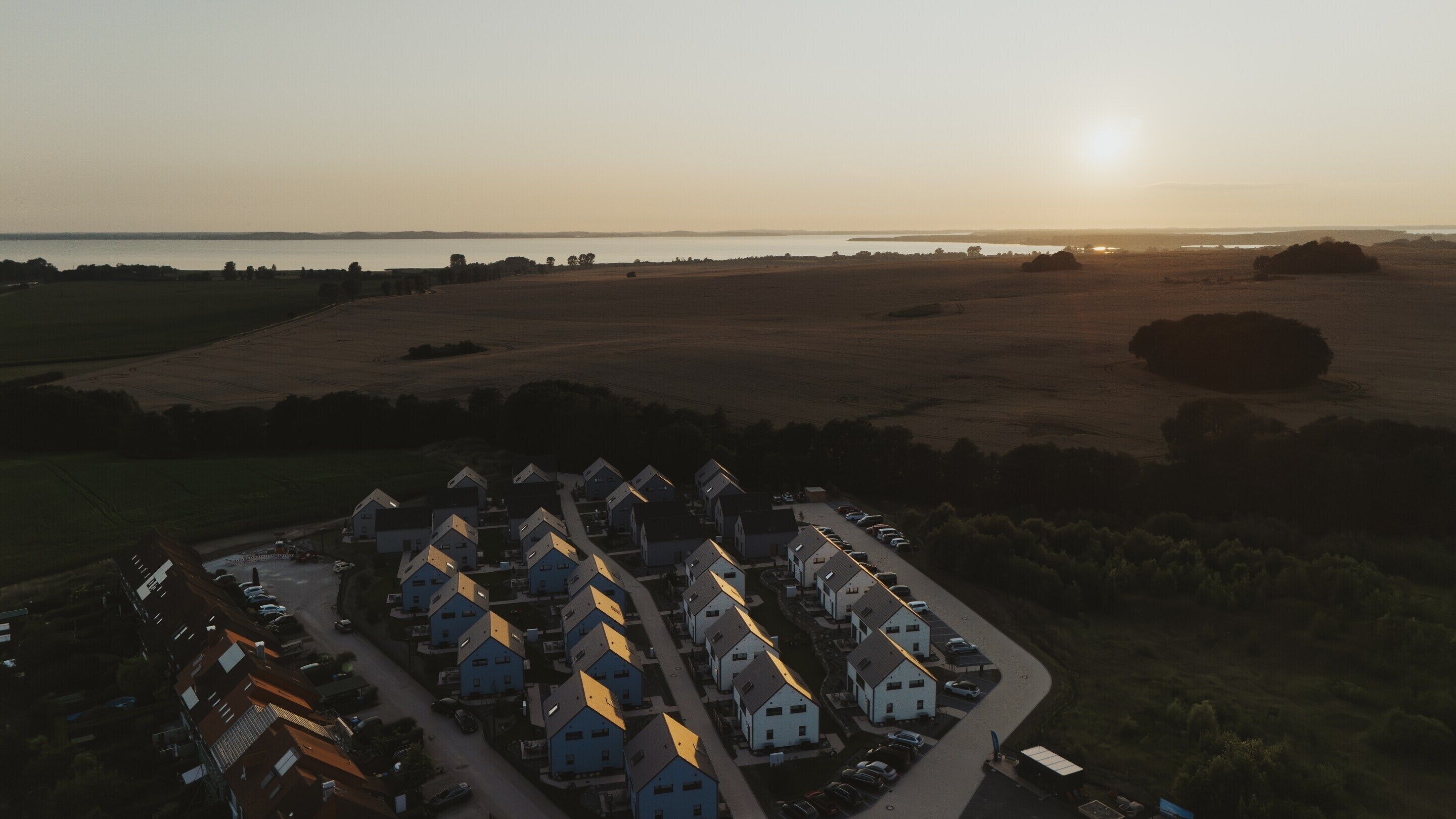 An aerial view of a residential area with houses arranged in rows, a parking lot, and a body of water in the background.