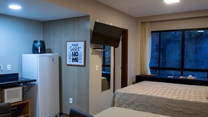 Hotellihuone, jossa on sänky, työpöytä tietokoneella, jääkaappi, ikkuna, josta on näkymä, ja ovi, joka johtaa toiseen huoneeseen.