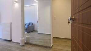Um corredor com porta de madeira, parede branca e um sofá cinzento ao fundo.
