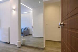 A hallway with a wooden door, a white wall, and a grey sofa in the background. - Icarus Minimal 2bedroom Bliss (Athina)