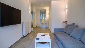 Uma sala de estar moderna com uma televisão de tela plana, um sofá cinzento, uma mesa de centro branca e uma cozinha ao fundo.