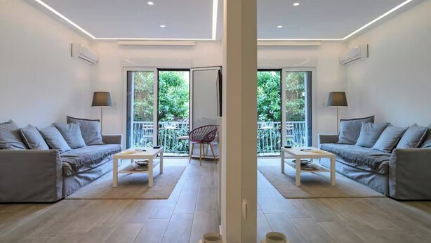 Uma sala de estar moderna com dois sofás, uma mesa de centro e uma poltrona. Janelas amplas e uma varanda são visíveis.