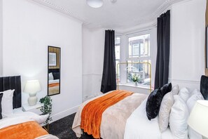 A hotel room with a large bed, a bedside table with a lamp, a window with curtains, and a view of the city. - 3BR House | 15 min Walk to Anfield | Free Parking (Liverpool)