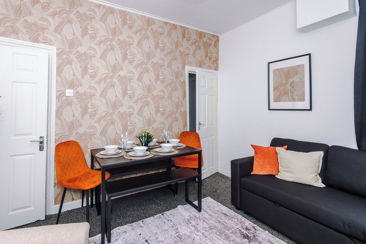 A dining area with a table set for four, two chairs, a black sofa with cushions, and a framed picture on the wall.