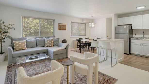 Ein modernes Wohnzimmer mit Sofa, Couchtisch und Sesseln, ein Essbereich mit Tisch und Stühlen sowie eine Küche mit Schränken und Geräten.