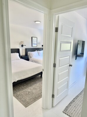 A hotel room with two beds, a nightstand, a lamp, a framed picture, and a door leading to another room. - Family Suite King Queen Queen + Queen sofa bed / Free Parking (Arlington)