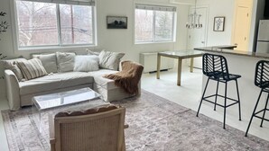 Ein modernes Wohnzimmer mit einer Couch, einem Couchtisch und Sesseln, ein Essbereich mit Tisch und Stühlen sowie eine Küche mit Kühlschrank.
