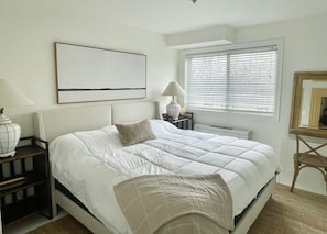 Une chambre à coucher comprenant un lit, des tables de chevet, une lampe, une fenêtre avec des stores et une œuvre d’art accrochée au mur.