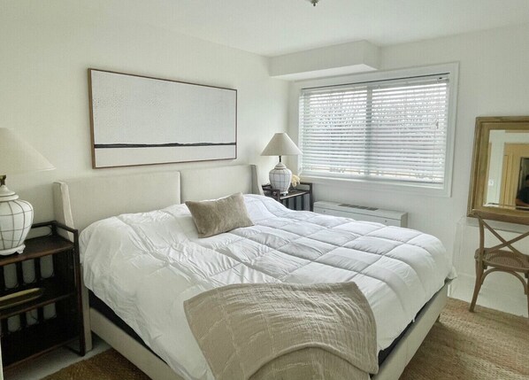 Une chambre à coucher comprenant un lit, des tables de chevet, une lampe, une fenêtre avec des stores et une œuvre d’art accrochée au mur.