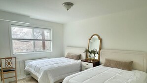 Ein Schlafzimmer mit zwei Betten, einem Holzstuhl, einem Nachttisch mit Spiegel und einem Fenster mit Jalousien.