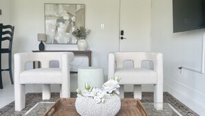 Una habitación con dos sillas blancas, una mesita de bandeja tejida, un jarrón con flores, una lámpara y un cuadro en la pared.