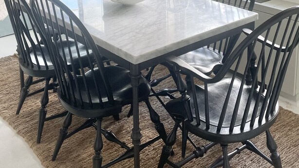 Un comedor con una mesa blanca, sillas negras, una lámpara colgante y vistas a árboles a través de ventanas amplias.