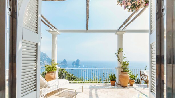 Un balcone con vista sul mare, arredi bianchi e piante in vaso.
