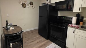 Angolo cottura compatto con frigorifero nero, microonde e forno, pavimento in legno e tavolo con sedie.