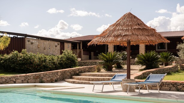 Una piscina con sillas de descanso y un paraguas de paja, rodeada de muros de piedra y follaje.