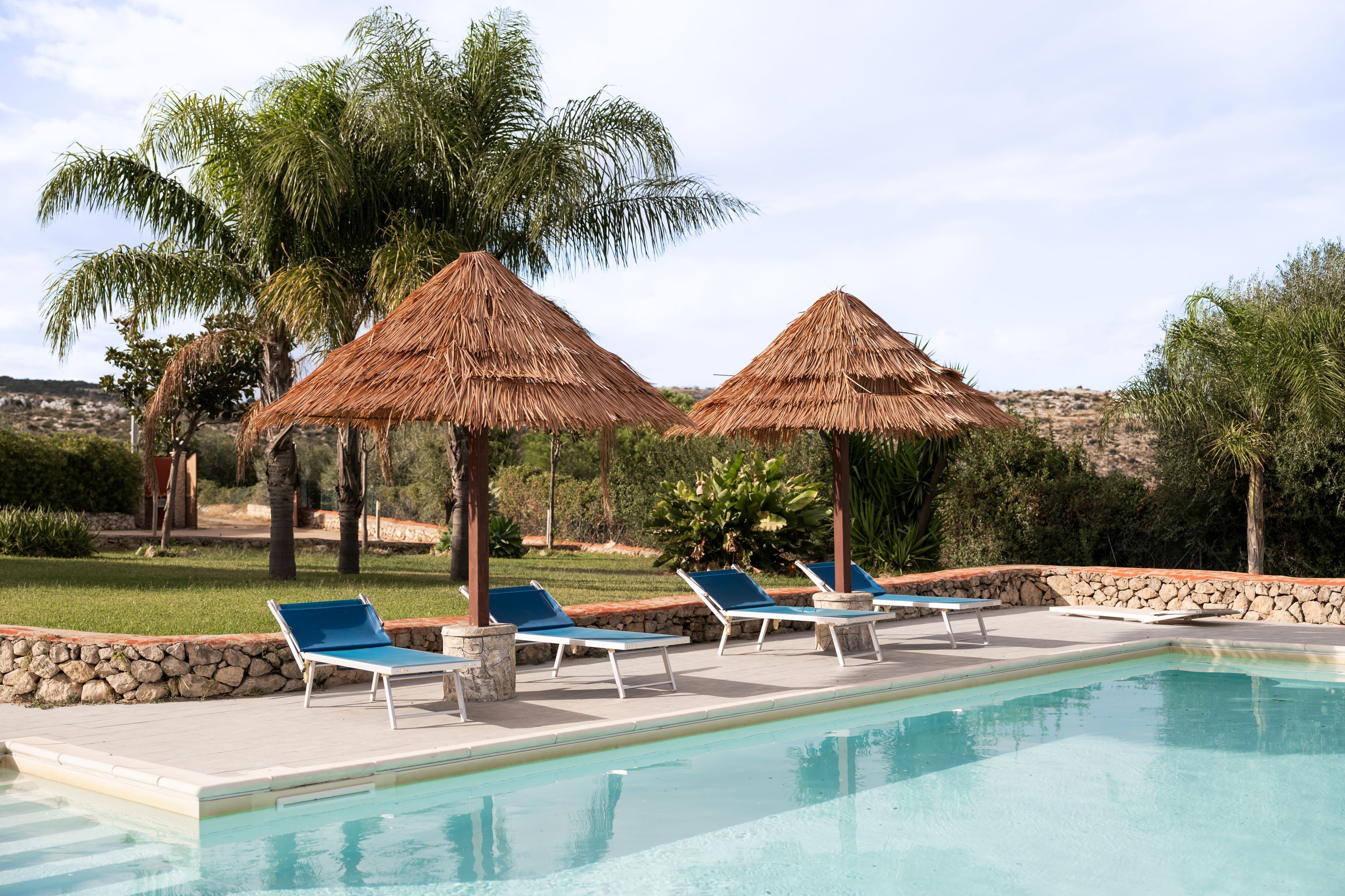 A pool area with lounge chairs and thatched umbrellas, surrounded by palm trees and a stone wall.