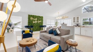 Ein modernes Wohnzimmer mit einer Couch, Sesseln, einem Couchtisch, einem Deckenventilator und einem Küchenbereich mit Schränken und einem Fenster.