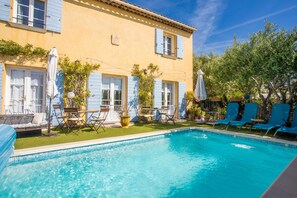 Un espace piscine avec une piscine bleue, des chaises longues et un bâtiment aux volets bleus.