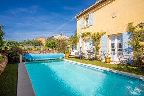 Une piscine bordée d’un mur en pierre et dotée d’une terrasse bleue, entourée d’une pelouse bien entretenue et d’un bâtiment jaune aux volets bleus.