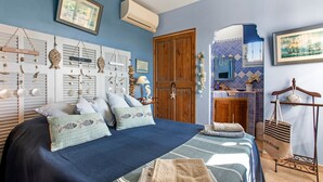 A bedroom with a bed, pillows, a hanging light fixture, a wooden door, and a bathroom visible through an archway. - Bed and Breakfast 'Chez Sophie - Chambre Côté Mer' with Mountain View, Wi-Fi and Air Conditioning (Roquebrune-sur-Argens)