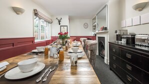 Ein Essbereich mit einem langen Holztisch, gedeckt für das Frühstück, einem Kamin und einer Küchenzeile mit Schränken und Geräten.