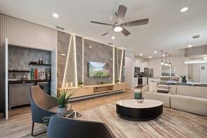 Una sala moderna con televisor de pantalla plana, ventilador de techo y comedor con cocina al fondo.